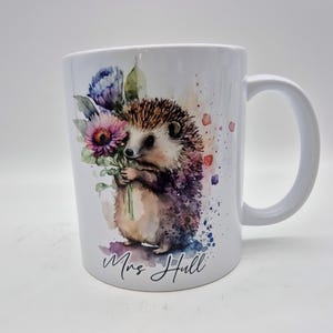 May include: White ceramic mug with a watercolour illustration of a hedgehog clutching flowers. The hedgehog is brown and tan with a spiky back. The flowers are purple and pink. The mug has the name "Mrs Hull" written on it.