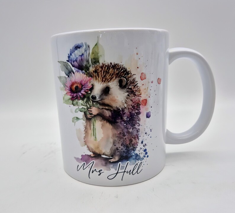 May include: White ceramic mug with a watercolour illustration of a hedgehog clutching flowers. The hedgehog is brown and tan with a spiky back. The flowers are purple and pink. The mug has the name "Mrs Hull" written on it.