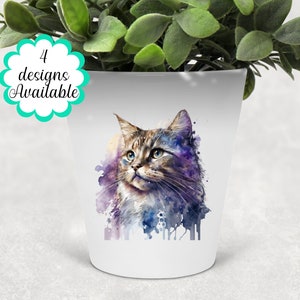 Könnte beinhalten: Ein weißer Keramik-Pflanztopf mit einem Aquarell-Design eines Katers mit blauen Augen. Der Kater ist von violetten und blauen Aquarell-Spritzen umgeben. Der Topf hat den Text "4 Designs verfügbar" auf der Vorderseite.