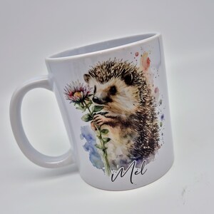 May include: White ceramic mug with a watercolour illustration of a hedgehog holding a flower. The hedgehog has brown and tan fur with a spiky back. The mug has a curved handle and the name "Mel" is written in cursive.