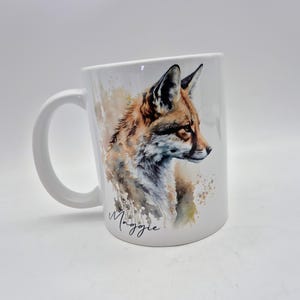 May include: White ceramic mug featuring a watercolor illustration of a fox in shades of brown, orange, and white. The mug has a curved handle and the name "Maggie" is written in cursive.