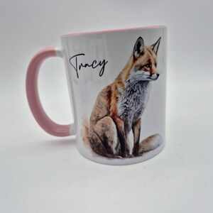 May include: White ceramic mug with a pink handle and rim. The mug features a watercolor illustration of a fox and the name "Tracy" in black script. The fox is depicted with orange, brown, and white fur.