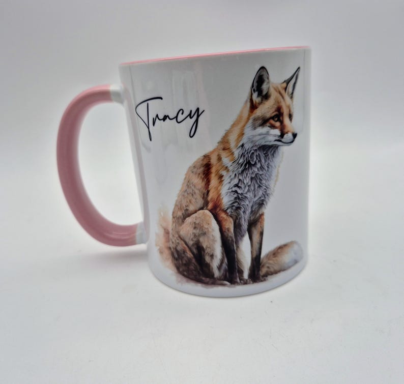 May include: White ceramic mug with a pink handle and rim. The mug features a watercolor illustration of a fox and the name "Tracy" in black script. The fox is depicted with orange, brown, and white fur.