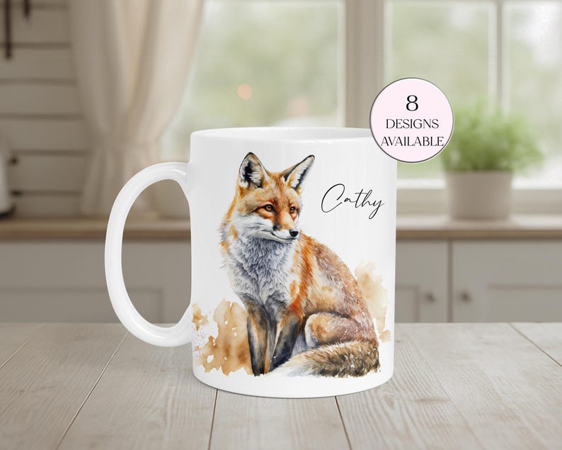 May include: White ceramic mug featuring a watercolor illustration of a fox with orange, brown, and gray fur. The mug has a curved handle and the name "Cathy" is written on the side. A pink circle indicates that 8 designs are available.