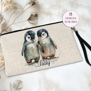 May include: A rectangular beige clutch bag with a black zip and wrist strap. The clutch features a watercolour illustration of two penguin chicks. The word "Tally" is written below the penguins, and the text "8 Designs Available" is in a pink circle.