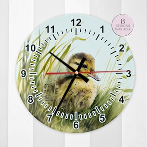 Könnte beinhalten: Runde Wanduhr mit einer Aquarellillustration eines Entenküken, das in hohem, grünem Gras sitzt. Das Zifferblatt hat schwarze Ziffern und Zeiger mit einem roten Sekundenzeiger. Der Text "8 DESIGNS AVAILABLE" befindet sich in einem rosa Kreis.