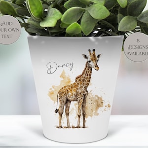 May include: White ceramic planter featuring a watercolor giraffe design with brown and tan spots. The planter has the name "Darcy" in script and the text "8 Designs Available". The planter is holding a green plant.
