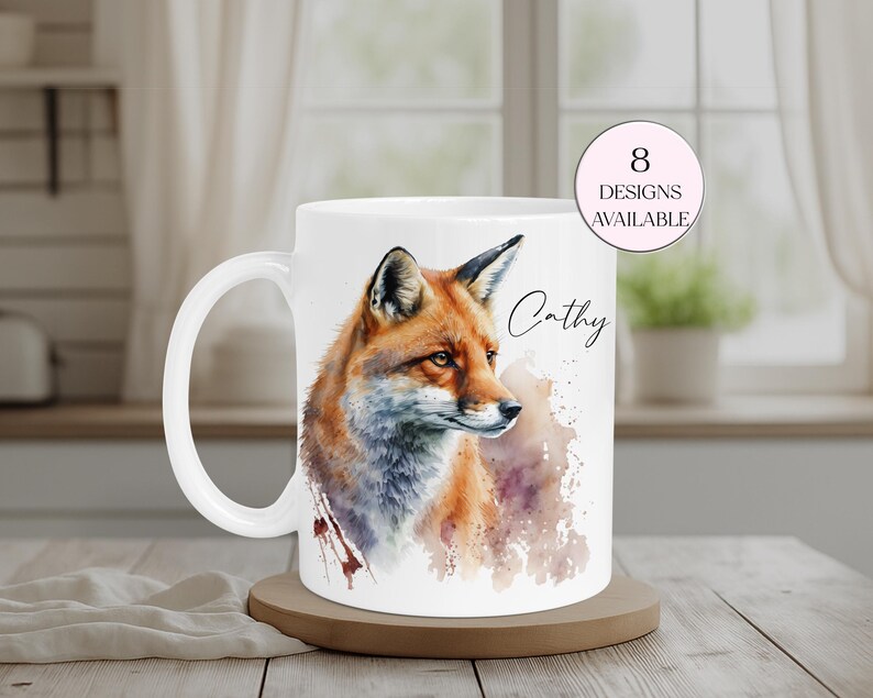 May include: White ceramic mug featuring a watercolor illustration of a fox with orange, brown, and blue tones. The mug has a white handle and the name "Cathy" is written on the side. A pink circle indicates "8 designs available".