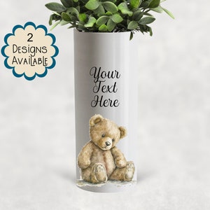 May include: A white cylindrical vase featuring a watercolour teddy bear illustration. The vase has the text "Your Text Here" and a label stating "2 Designs Available". The vase is filled with green plants.