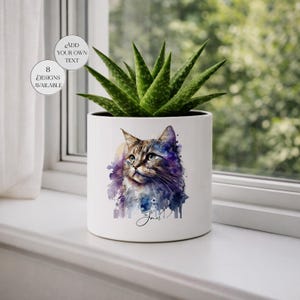Könnte beinhalten: Weißer Keramik-Pflanztopf mit einem Aquarellgemälde einer Katze mit blauen Augen und einem lila und blauen Hintergrund. Der Topf enthält eine grüne Aloe Vera-Pflanze. Auf dem Topf steht "Jan" und "Fügen Sie Ihren eigenen Text hinzu" und "8 Designs verfügbar".