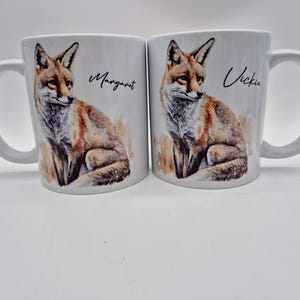 May include: Two white ceramic mugs with a watercolor illustration of a fox. One mug has the name "Margaret" and the other has "Vickie" written on it. The fox is depicted in shades of brown, orange, and white.