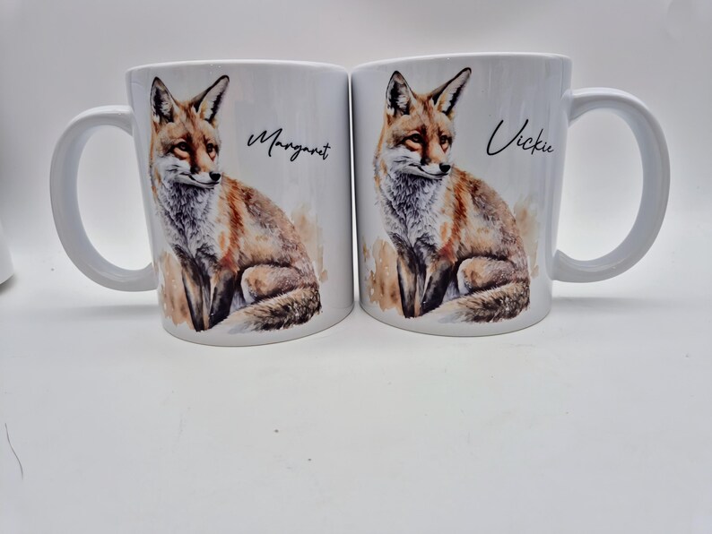 May include: Two white ceramic mugs with a watercolor illustration of a fox. One mug has the name "Margaret" and the other has "Vickie" written on it. The fox is depicted in shades of brown, orange, and white.