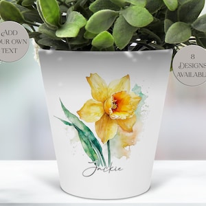 May include: White ceramic flower pot featuring a watercolor daffodil design with yellow petals and green leaves. The pot has the name "Jackie" written in script. The pot also has the text "Add your own text" and "8 designs available".
