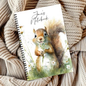 Könnte beinhalten: Spiralgebundenes Notizbuch mit einer Aquarellillustration eines lächelnden Eichhörnchens, das Wildblumen hält. Auf dem Notizbuchcover steht in handschriftlicher Schrift der Text "Jan's Notebook". Das Eichhörnchen ist braun und hellbraun, vor einem Hintergrund aus grünem Gras und weißen Blumen.