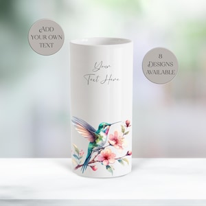 May include: A white cylindrical tumbler featuring a watercolor illustration of a hummingbird perched on a branch with pink flowers. The text "Your Text Here" is printed in black script. Two circular badges state "ADD YOUR OWN TEXT" and "8 DESIGNS AVAILABLE".