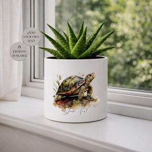May include: A white cylindrical ceramic planter containing a green succulent plant. The front of the planter features a watercolour illustration of a tortoise on a rock with the text "Your Text" below it. Two circular badges state "ADD YOUR OWN TEXT" and "8 DESIGNS AVAILABLE".