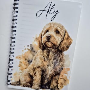 May include: A spiral-bound notebook with a watercolour illustration of a light brown dog. The dog is depicted sitting, with a fluffy coat and expressive eyes. The name "Aly" is written in cursive at the top. Brown and tan watercolour splashes add a decorative element.