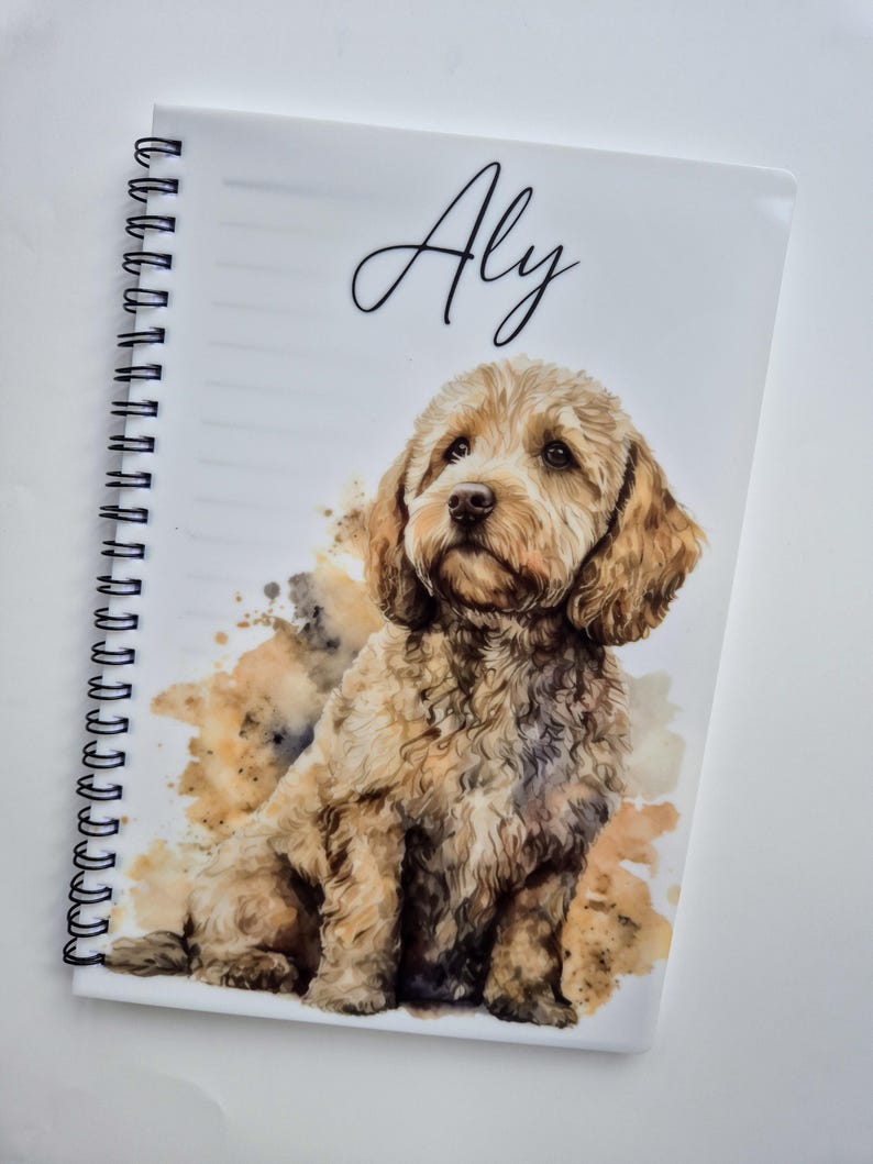 May include: A spiral-bound notebook with a watercolour illustration of a light brown dog. The dog is depicted sitting, with a fluffy coat and expressive eyes. The name "Aly" is written in cursive at the top. Brown and tan watercolour splashes add a decorative element.