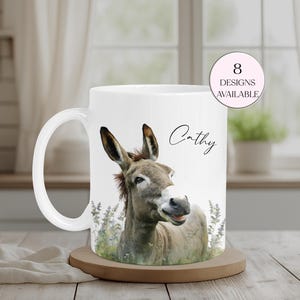 May include: White ceramic mug with a donkey illustration. The mug has a curved handle and the name "Cathy" is written on the side. A pink circle indicates "8 designs available". The mug sits on a wooden coaster. The mug is suitable for approximately 300 ml of liquid.
