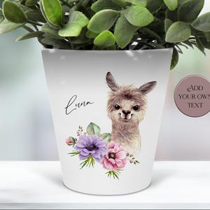 May include: White ceramic plant pot decorated with a watercolour illustration of a llama and a floral arrangement of purple and pink flowers. The name "Luna" is written in cursive. A button on the side reads "Add Your Own Text".