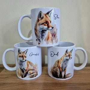 May include: Three white ceramic mugs with fox illustrations. Each mug features a different fox portrait and the words "She", "Grandad", and "Dad" in black script. The mugs are stacked on a wooden surface.