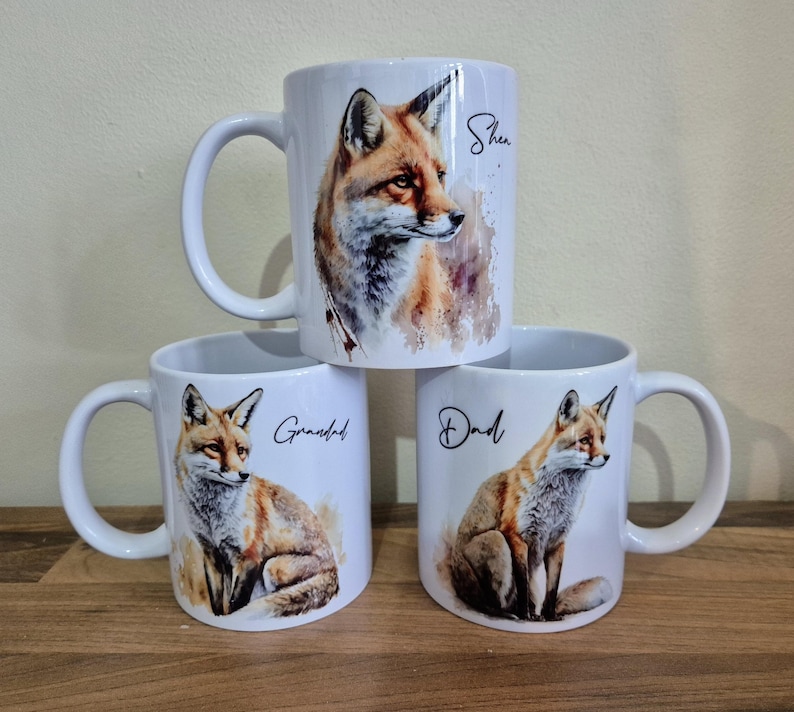 May include: Three white ceramic mugs with fox illustrations. Each mug features a different fox portrait and the words "She", "Grandad", and "Dad" in black script. The mugs are stacked on a wooden surface.