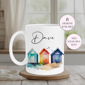 May include: White ceramic mug with a watercolour design of three beach huts in turquoise, orange, and blue. The mug has the name "Dave" in cursive and the text "8 designs available" and "Add your own text".