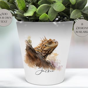 May include: White ceramic plant pot with a watercolour illustration of an iguana on a log. The pot has the name "Jackie" in script and the text "Add Your Own Text" and "8 Designs Available" in circles.