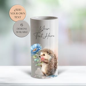 May include: A cylindrical, light grey tumbler featuring a watercolour illustration of a hedgehog holding a flower. The tumbler has the text "Your Text Here" and two circular text labels: "Add Your Own Text" and "6 Designs Available."