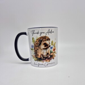 May include: White ceramic mug with a navy blue handle and rim. The mug features a watercolour illustration of a hedgehog surrounded by flowers. Text on the mug reads "Thank you Aiden" and "love from Grace x".