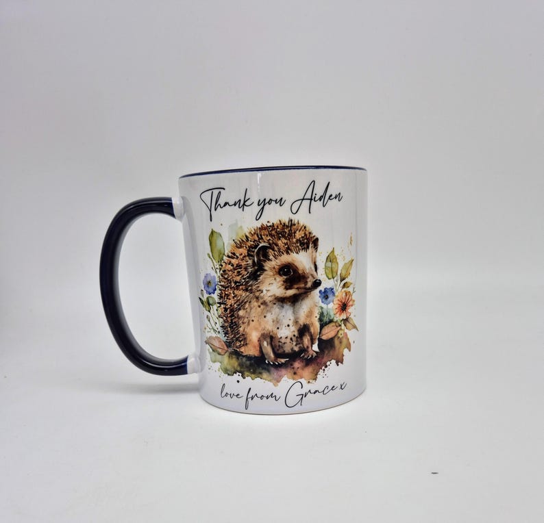 May include: White ceramic mug with a navy blue handle and rim. The mug features a watercolour illustration of a hedgehog surrounded by flowers. Text on the mug reads "Thank you Aiden" and "love from Grace x".