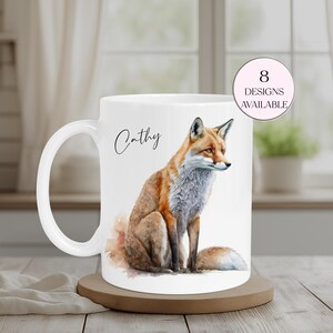 May include: White ceramic mug featuring a watercolor illustration of a fox with orange, brown, and gray fur. The mug has the name "Cathy" in script and a pink circle with the text "8 Designs Available". The mug sits on a wooden coaster.