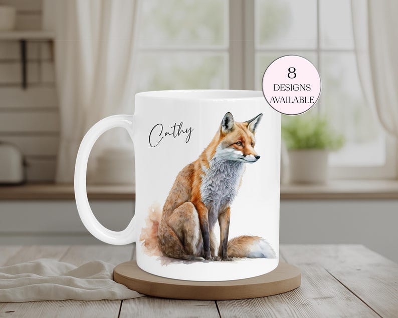 May include: White ceramic mug featuring a watercolor illustration of a fox with orange, brown, and gray fur. The mug has the name "Cathy" in script and a pink circle with the text "8 Designs Available". The mug sits on a wooden coaster.