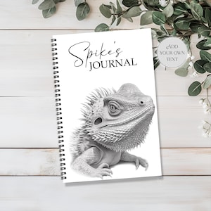 May include: A spiral-bound journal featuring a black and white lizard illustration. The cover displays the text "Spike's JOURNAL" in a script font. The journal rests on a white wooden surface, with greenery subtly placed in the background.