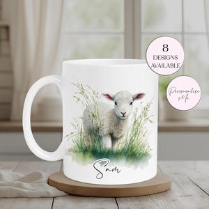 May include: White ceramic mug with a watercolour illustration of a lamb in long grass. The mug has a curved handle and the name "Sam" is written in script. Two pink circles with text are also visible.