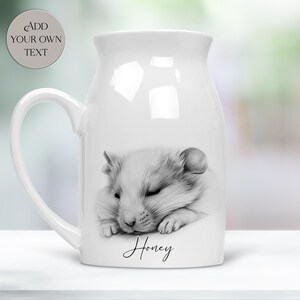 May include: A white ceramic mug featuring a black and white sketch of a sleeping hamster. Below the hamster, the name Honey is printed in black script. A circular badge in the upper left corner contains the text ADD YOUR OWN TEXT.