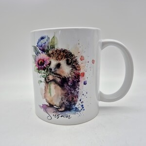 May include: White ceramic mug with a watercolour illustration of a hedgehog holding flowers. The hedgehog has brown and purple fur, with a light brown body. The mug has the name "Susan" written in cursive.