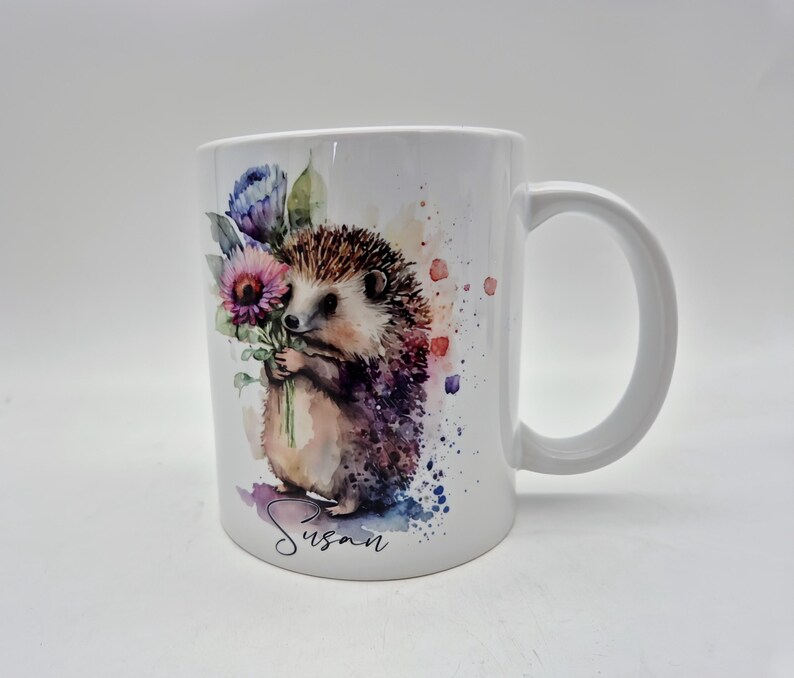 May include: White ceramic mug with a watercolour illustration of a hedgehog holding flowers. The hedgehog has brown and purple fur, with a light brown body. The mug has the name "Susan" written in cursive.