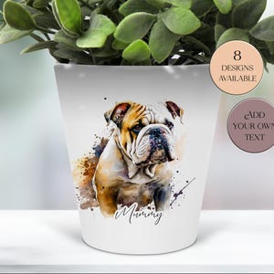 Könnte beinhalten: Weißer Keramik-Blumentopf mit einer Aquarell-Illustration einer Bulldogge. Der Hund hat braunes und weißes Fell, darunter steht das Wort "Mummy". Der Topf ist mit grünen Pflanzen gefüllt. Auf dem Topf steht "8 Designs verfügbar" und "Fügen Sie Ihren eigenen Text hinzu".