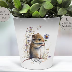May include: White ceramic plant pot featuring a watercolor illustration of a mouse holding a blue flower. The pot has the name "Tilly" written on it. The pot also has the text "Add Your Own Text" and "8 Designs Available".