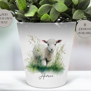 以下が含まれることがあります： 白いセラミック植木鉢。草むらにいる子羊の水彩イラストが描かれています。植木鉢には「Aimee」という名前が筆記体で書かれています。「Add Your Own Text」と「8 Designs Available」の文字も表示されています。