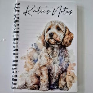 May include: A notebook with a spiral binding, featuring the text "Katie's Notes". The cover displays a watercolour illustration of a dog, predominantly light brown and white. The dog is depicted sitting, with a soft, neutral background.