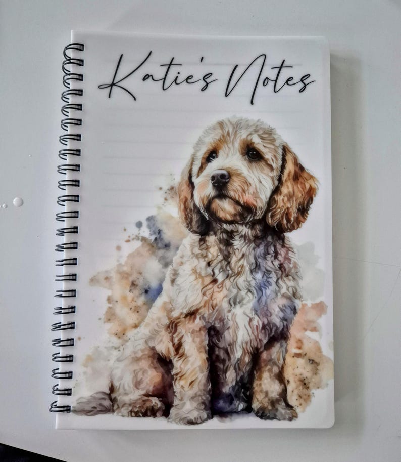 May include: A notebook with a spiral binding, featuring the text "Katie's Notes". The cover displays a watercolour illustration of a dog, predominantly light brown and white. The dog is depicted sitting, with a soft, neutral background.