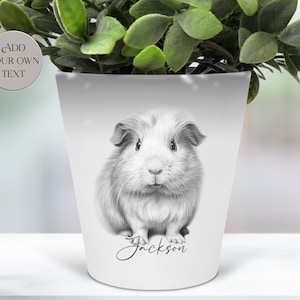 Peut inclure: Pot de fleurs en céramique blanche avec une illustration en niveaux de gris d'un cochon d'Inde. Le cochon d'Inde est au centre, avec le nom "Jackson" en cursive en dessous. Le pot contient des plantes vertes.