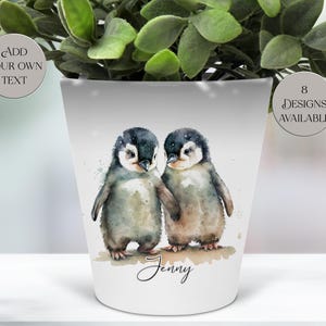 May include: White ceramic plant pot featuring a watercolour illustration of two penguins. The penguins are holding hands against a snowy backdrop. The name "Jenny" is written below. The pot has the text "Add your own text" and "8 designs available".