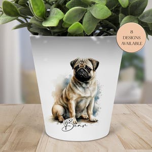 May include: White ceramic plant pot with a watercolour-style pug illustration. The pug is tan and black, with the name "Bear" below. A sticker indicates "8 Designs Available". The pot contains a green plant.