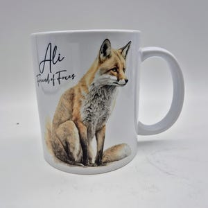 May include: White ceramic mug featuring a watercolor illustration of a fox in shades of brown, tan, and white. The mug has the text "Ali Friend of Foxes" in black script. The mug has a large, curved handle.