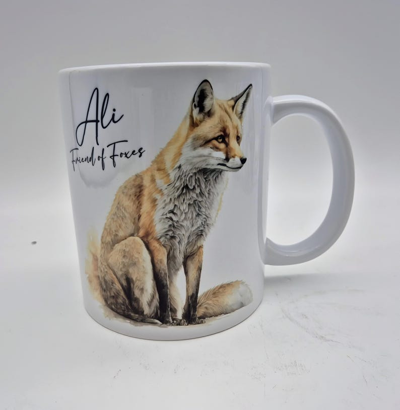 May include: White ceramic mug featuring a watercolor illustration of a fox in shades of brown, tan, and white. The mug has the text "Ali Friend of Foxes" in black script. The mug has a large, curved handle.