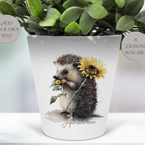 May include: White ceramic flower pot with a watercolour illustration of a hedgehog holding a sunflower. The pot has the text "Aimee" and the text "Add Your Own Text" and "8 Designs Available". The pot is filled with green leafy plants.