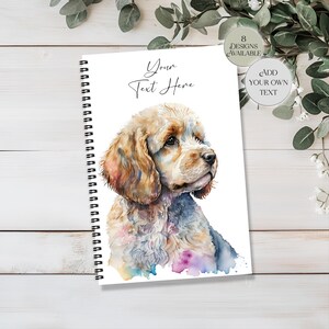May include: A spiral-bound notebook with a watercolour illustration of a Cocker Spaniel. The dog has tan and white fur with colourful accents. The notebook cover has the text "Your Text Here" and design options.